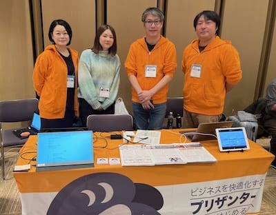 エコー電子工業株式会社様を訪問したときの写真。プリザンターの展示ブースの前に 4 人が並んでいる
