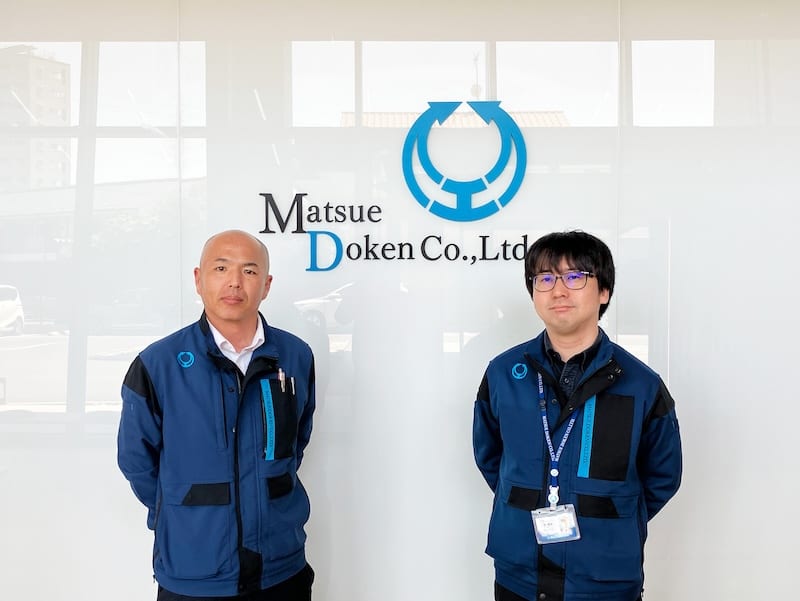Matsue Doken Co.,Ltd のロゴの前に立つ、松江土建株式会社のご担当者 2 名の写真