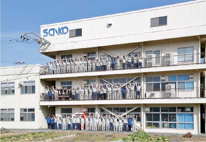 SANKO の社屋のベランダと前庭に並び、両手を挙げる社員の皆さんの写真